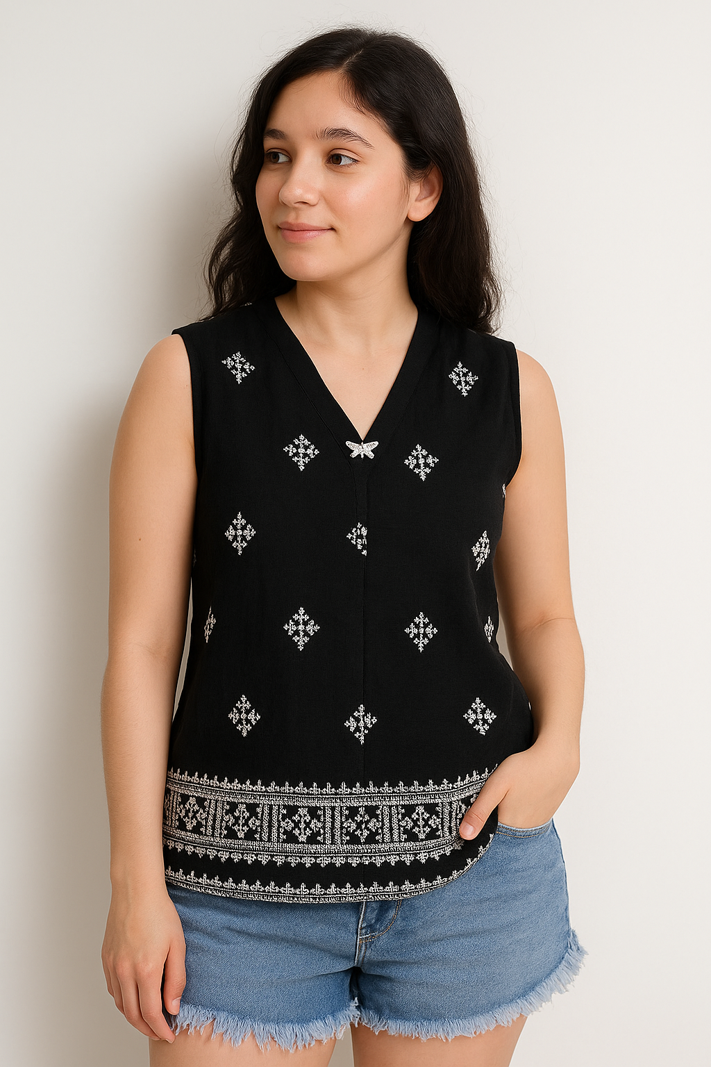 Woman wearing a black sleeveless top with white patterns and denim shorts on a plain background